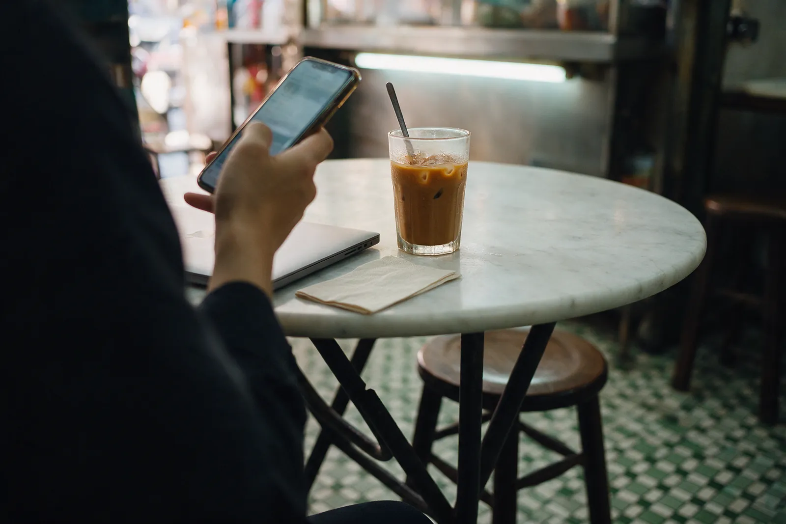 A person checking a phone at a marble kopitiam table, next to a closed laptop and an iced drink.