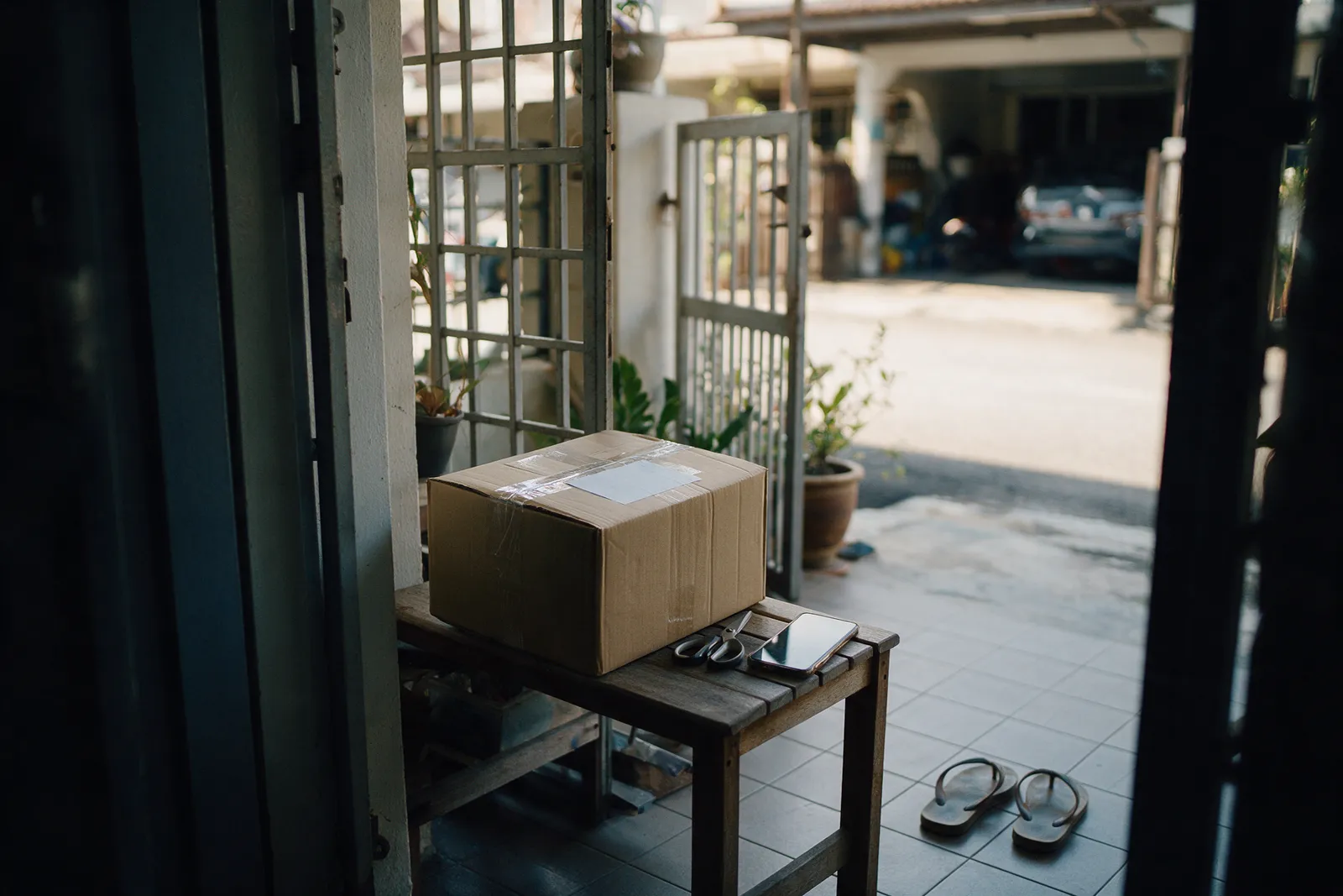 A plain brown courier box sealed with clear tape on a wooden bench in the shaded porch of a Malaysian terrace house, with scissors and a phone beside it