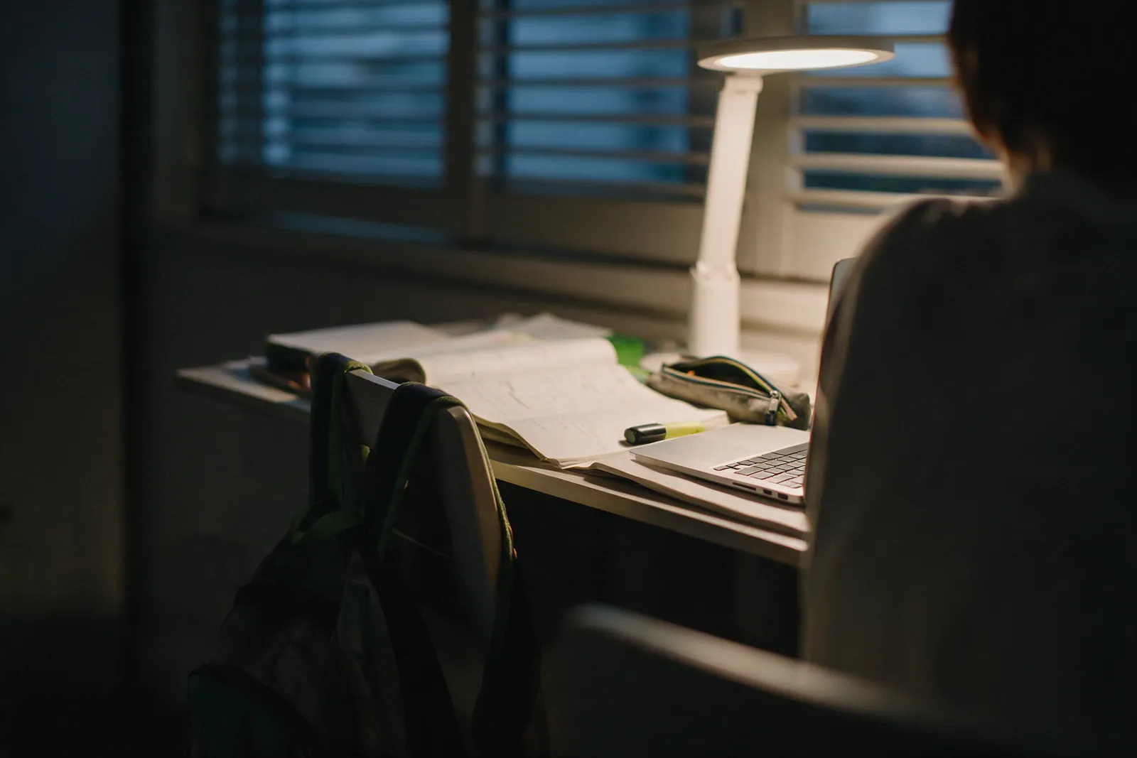 A student at a home study desk in the evening with a laptop, exercise books and a desk lamp.