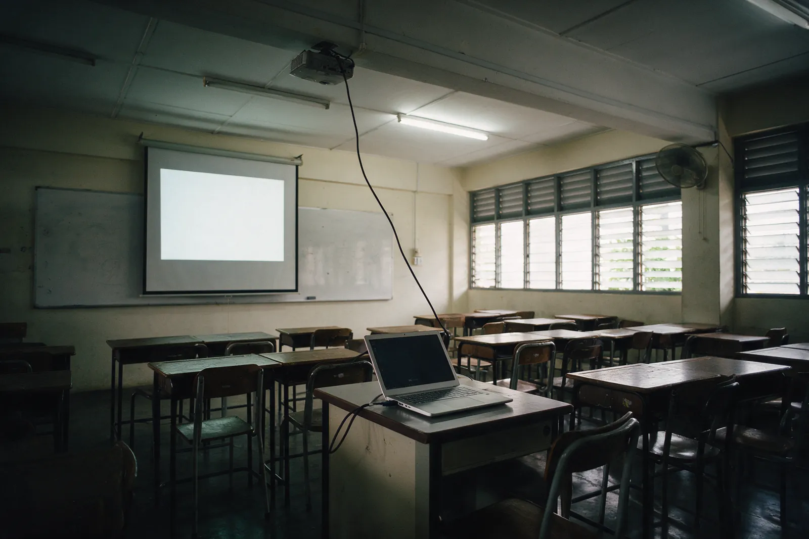An empty classroom with a laptop on the teacher's table cabled to a ceiling projector and screen.