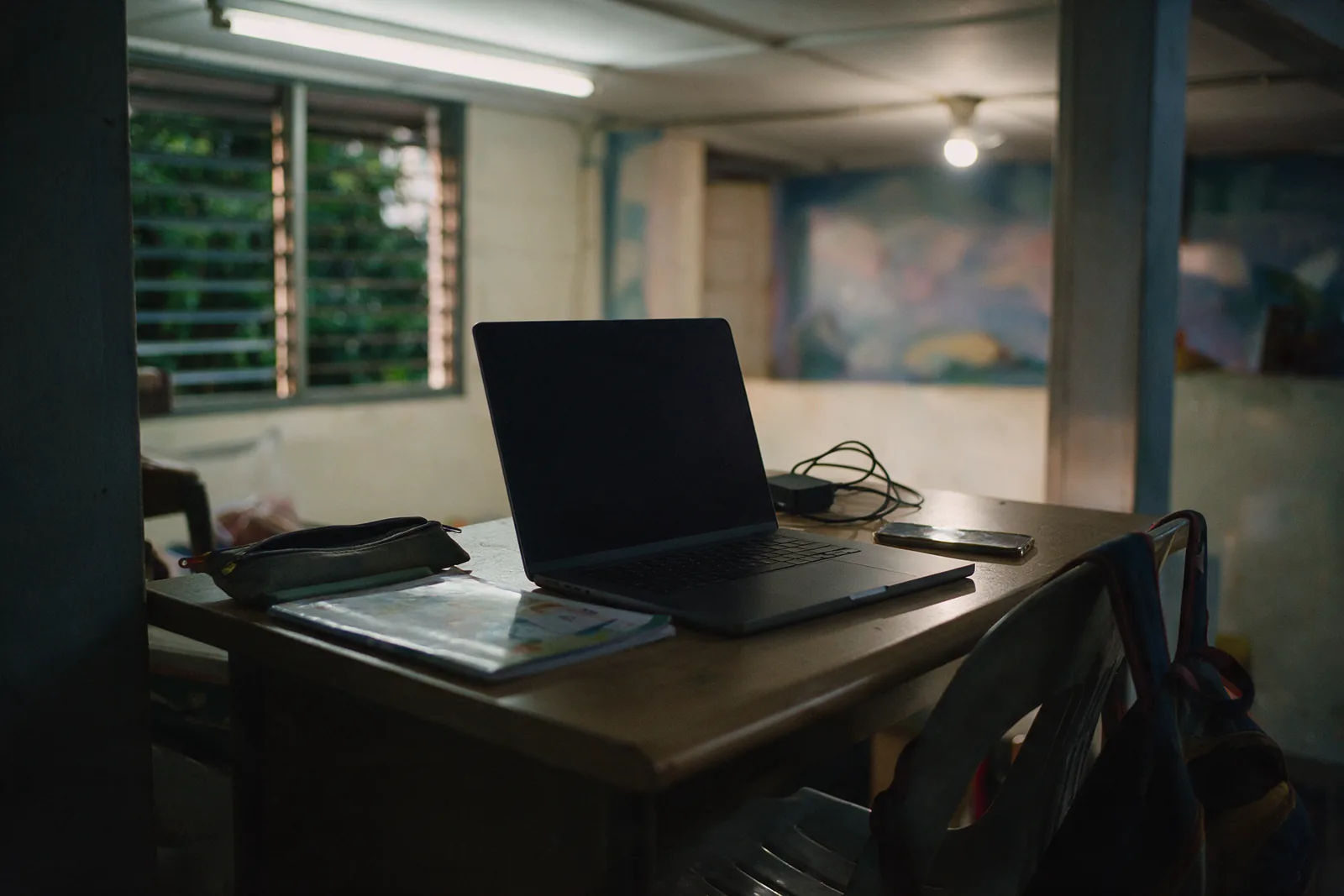 A laptop, a school exercise book and a pencil case on a study desk at home in the evening, a school bag on the chair behind.