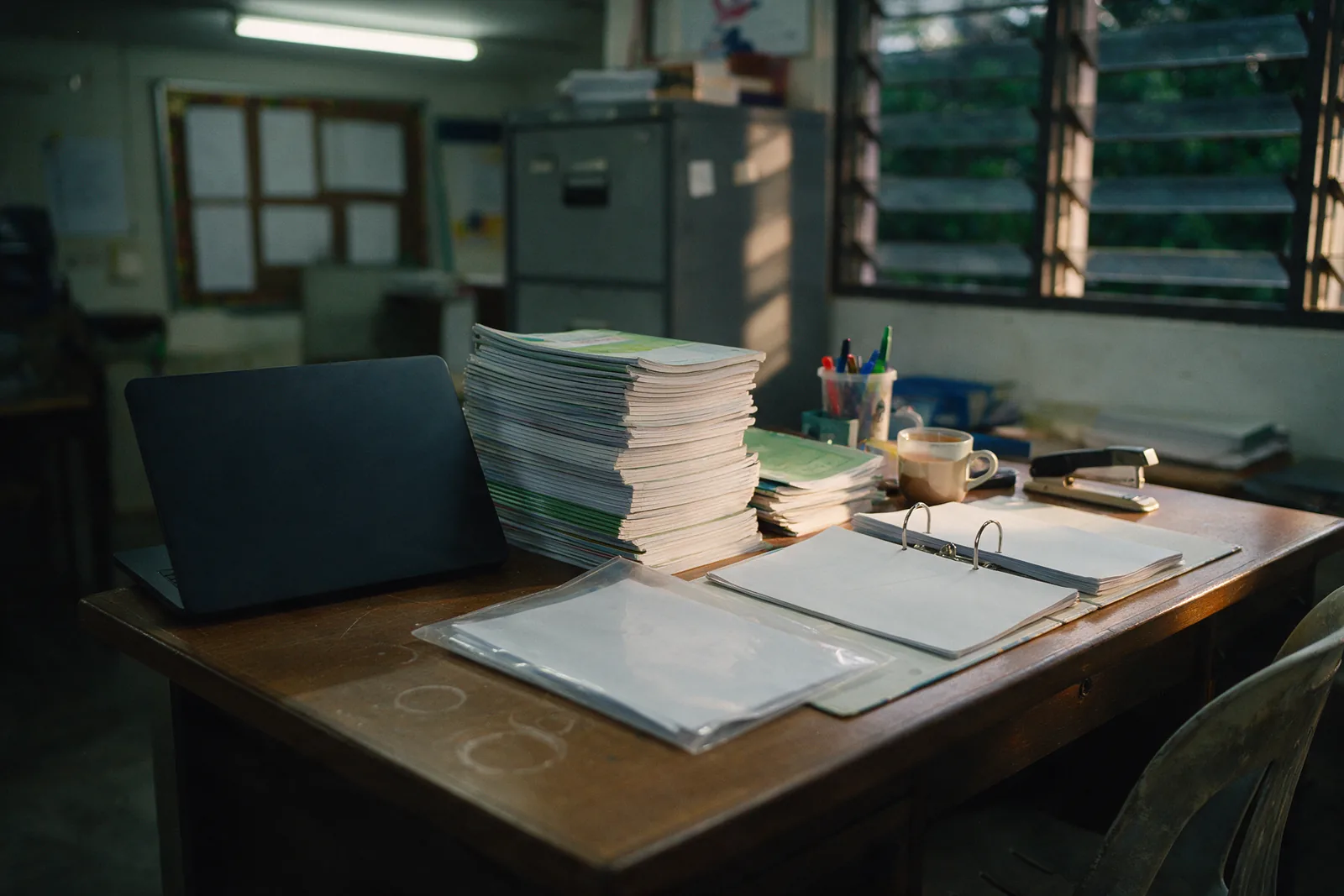 A teacher's staffroom desk in the late afternoon: an open plain laptop seen from behind, a tall stack of exercise books, an open ring binder and a mug of tea.